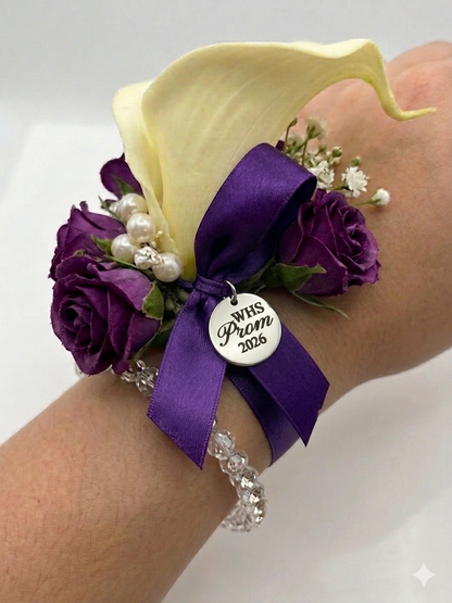 Wrist corsage with purple flowers, yellow calla lily, and 'WHS Prom 2023' charm on a wrist against a white background.