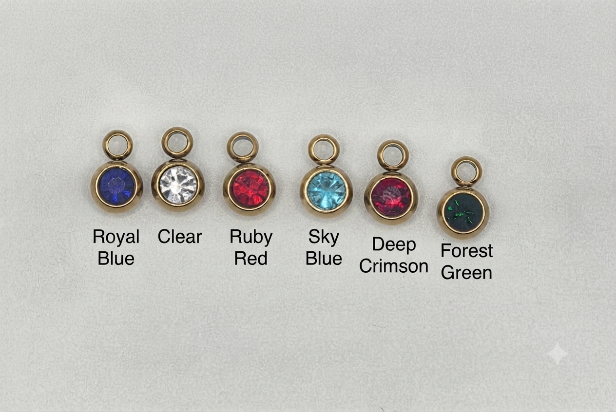 Six small oval gemstone charms in gold-toned bezels are arranged on a light gray background with printed color names underneath: Royal Blue, Clear, Ruby Red, Sky Blue, Deep Crimson, and Forest Green.