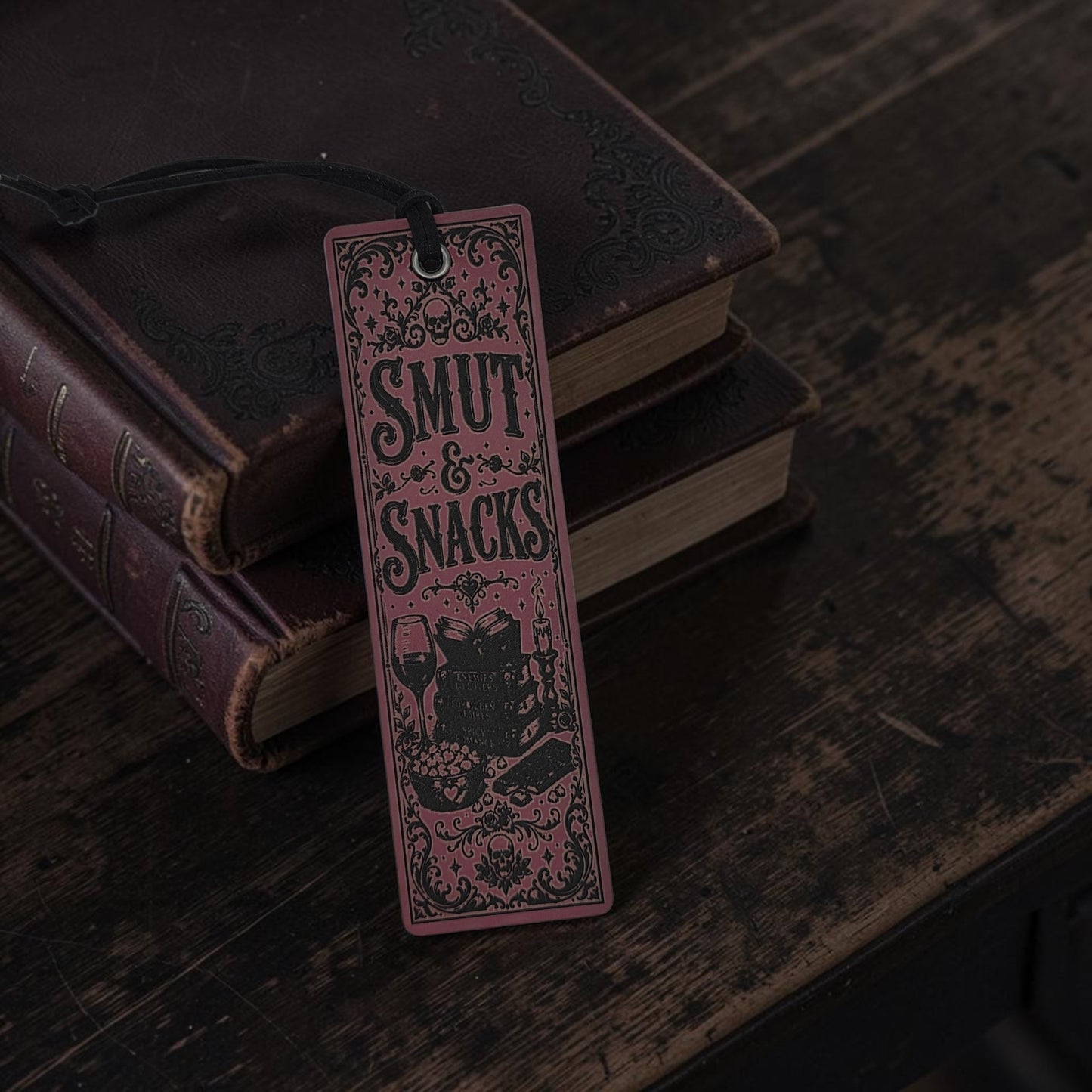 A rectangular bookmark or tag with a black hanging string, featuring the words “SMUT & SNACKS” in decorative lettering above an illustration of a cat, set against a stack of old books on a dark wooden surface.