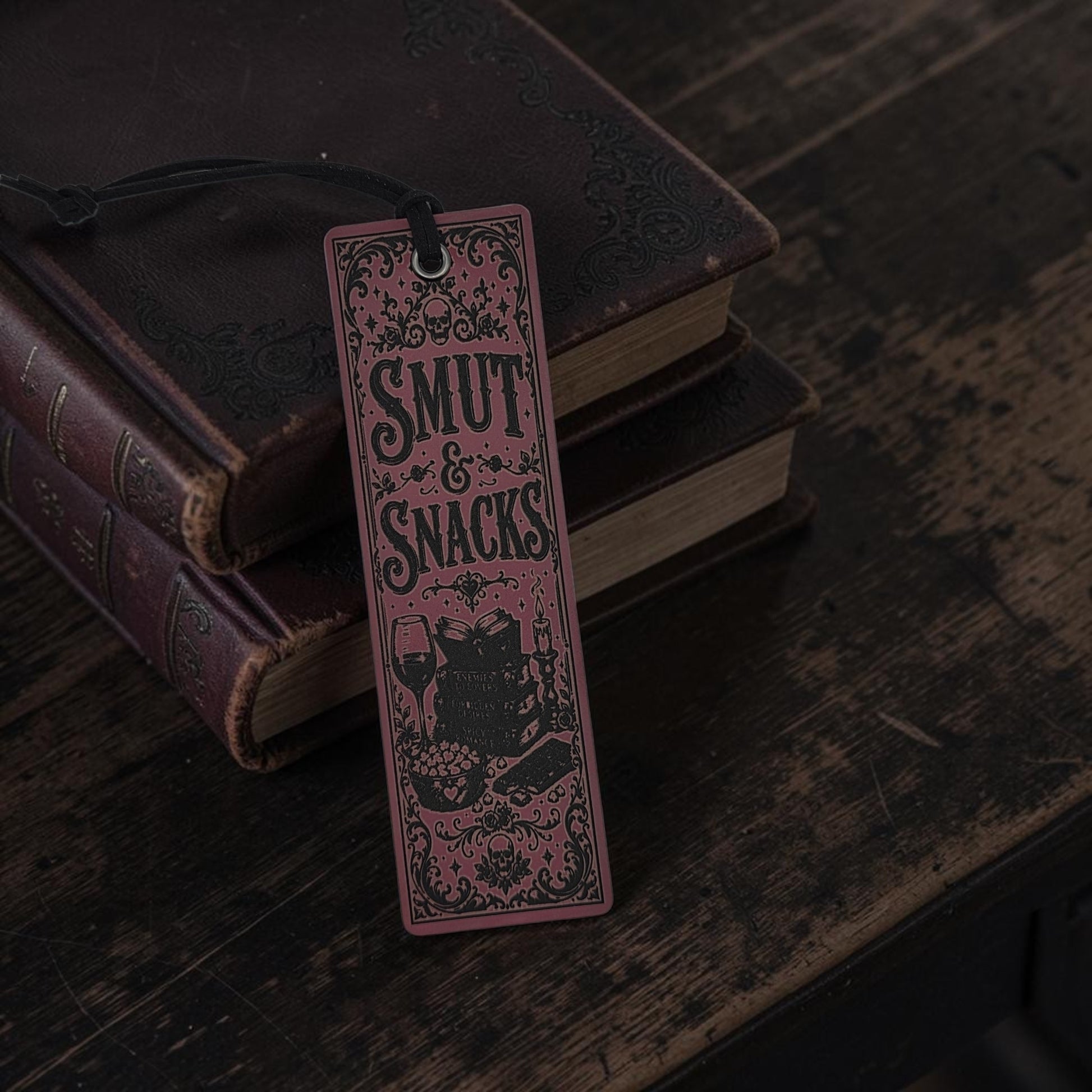 A rectangular bookmark or tag with a black hanging string, featuring the words “SMUT & SNACKS” in decorative lettering above an illustration of a cat, set against a stack of old books on a dark wooden surface.