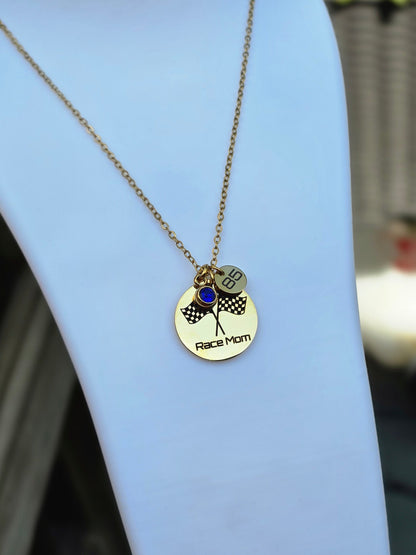 A gold-toned necklace displayed on a white jewelry bust. The pendant is a round disc engraved with 'Race Mom' and a checkered flag motif, with two small hanging charms, one engraved with '47'.
