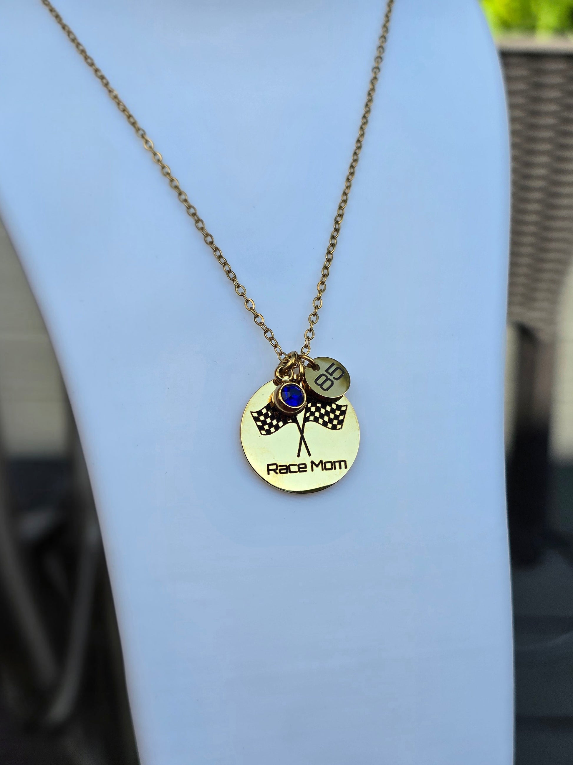 A gold-tone necklace displayed on a white bust, featuring a round pendant engraved with 'Race Mom' and two small charms, one with a blue stone and one with a checkered flag design.