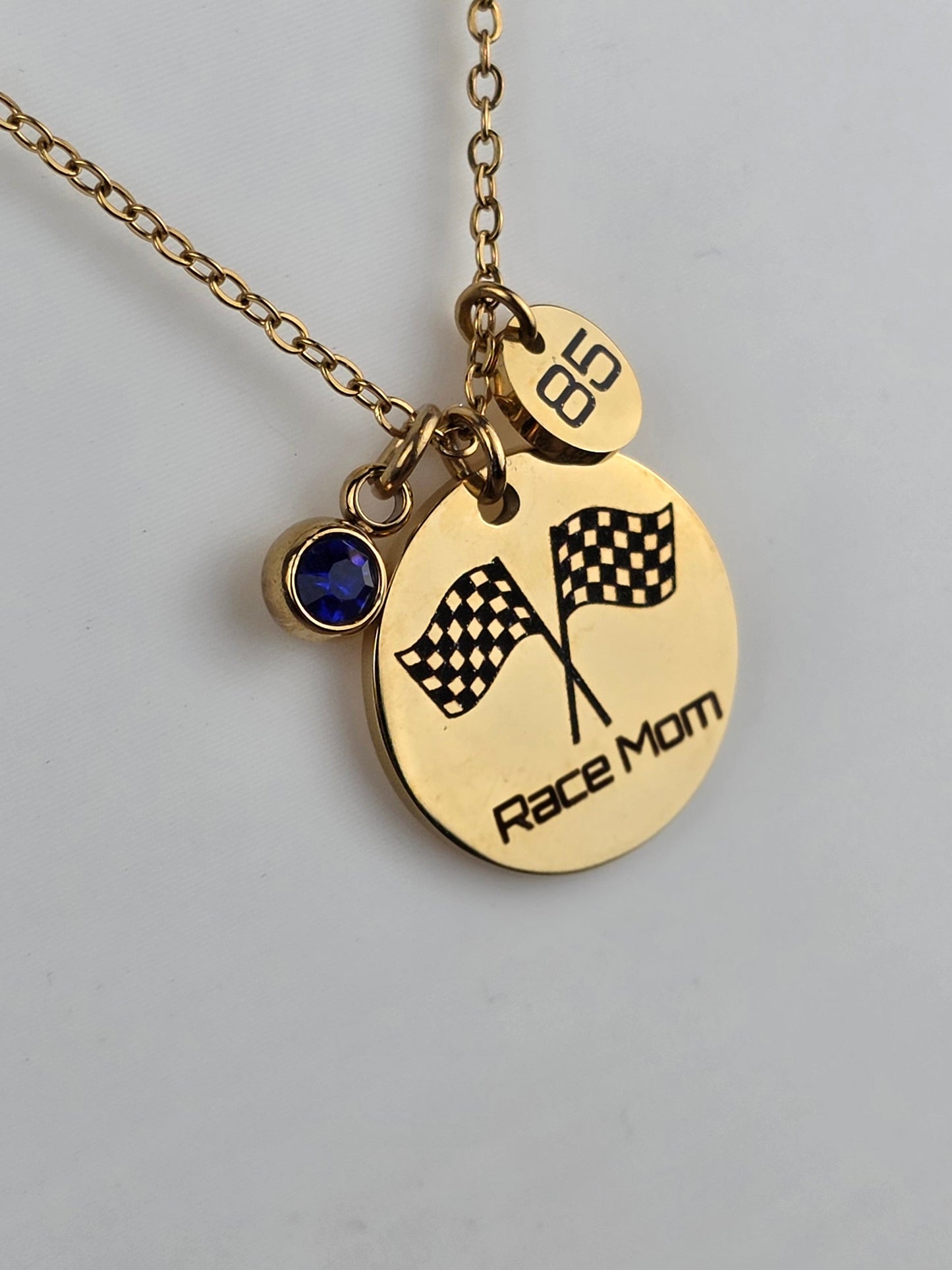 A gold-tone necklace with a round disc pendant engraved with 'Race Mom' and two checkered flags, plus a small round charm marked '68' and a dark blue gemstone charm, shown on a light gray background.