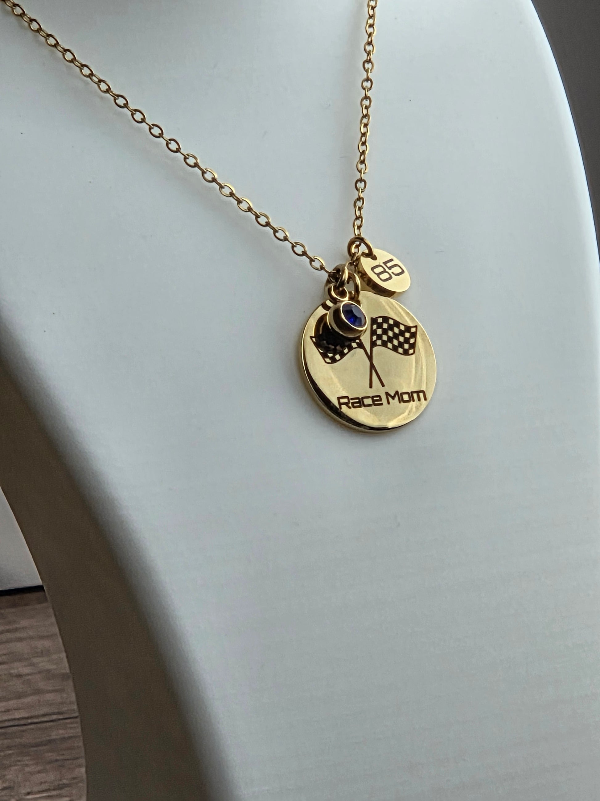 A gold chain necklace with a round 'Race Mom' pendant and a small heart charm, displayed on a white jewelry bust.
