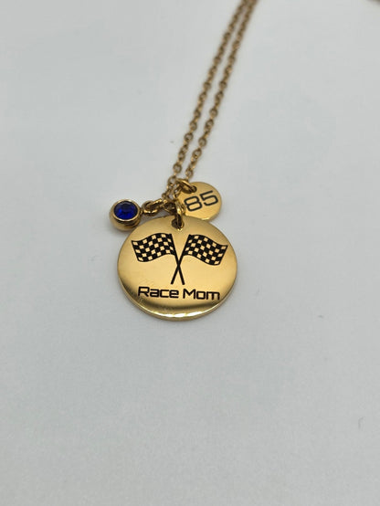 A gold necklace with a round pendant engraved with 'Race Mom' and two checkered flags, featuring a small heart charm and a tiny dark blue stone accent, displayed on a plain light gray background.