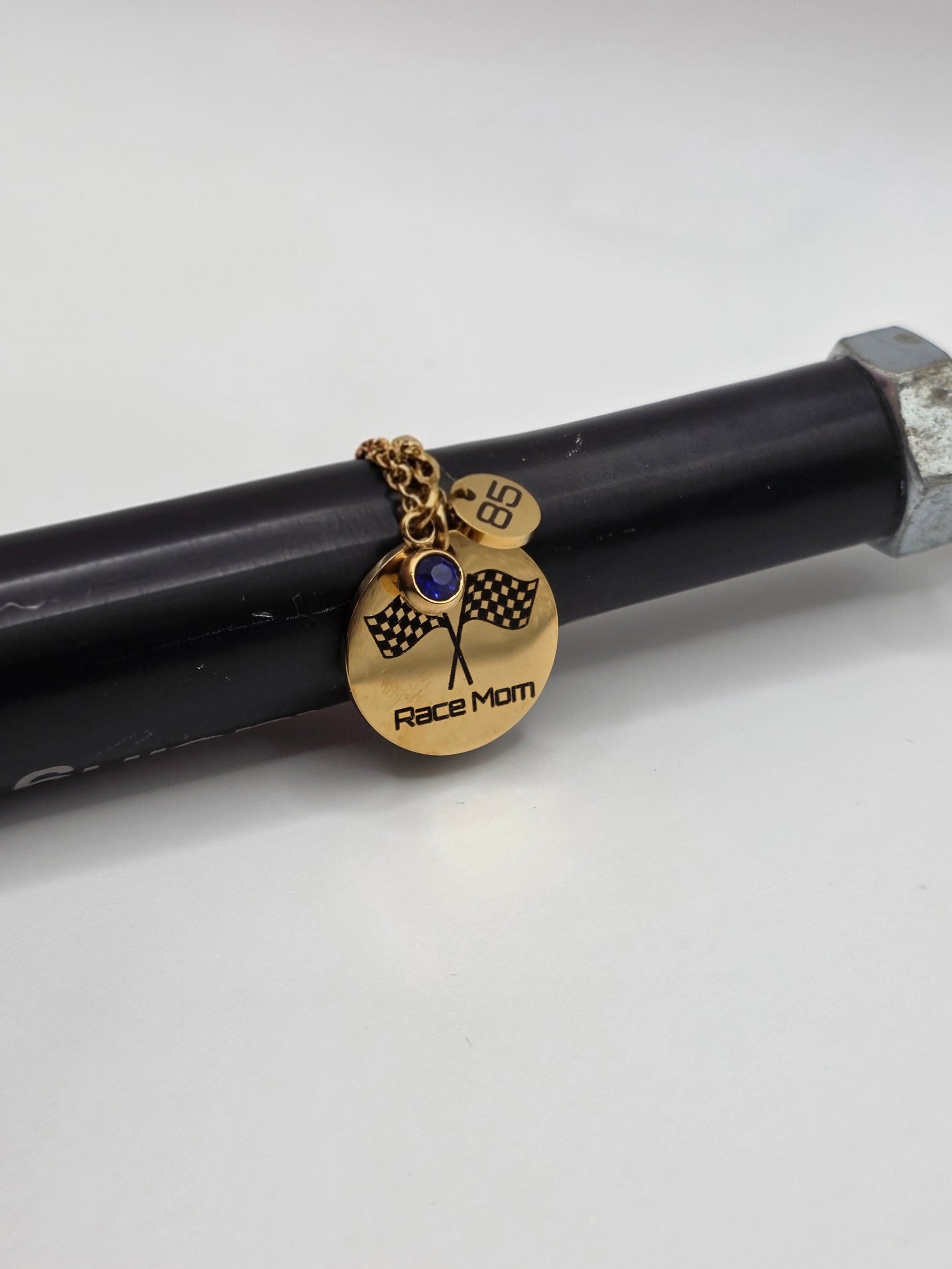 A gold-toned 'Race Mom' pendant charm with a blue stone and checkered flag design, shown hanging from a black rod on a white background.