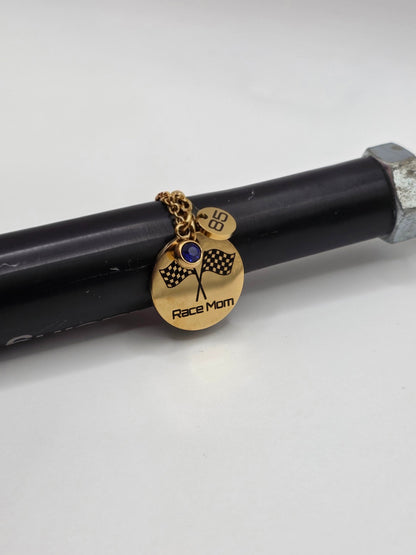 A gold-toned 'Race Mom' pendant charm with a blue stone and checkered flag design, shown hanging from a black rod on a white background.