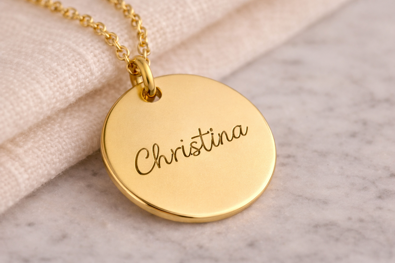 Gold necklace with a round pendant engraved with 'Christina' on a marble surface.