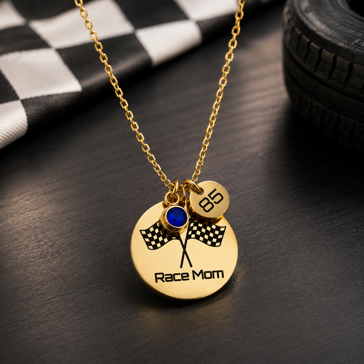 A gold-tone necklace with a round pendant engraved 'Race Mom' and crossed checkered flags, plus a small charm marked '85' and a blue gemstone charm, displayed on a dark surface with racing-themed props.