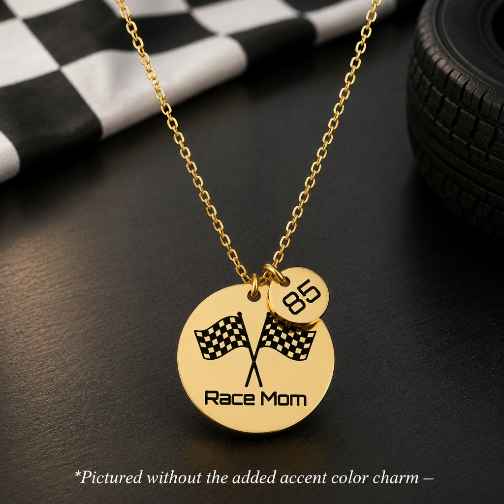 A gold chain necklace with a round gold pendant engraved with crossed checkered flags and the words 'Race Mom,' plus a small dangling charm marked '85.' The necklace is photographed on a dark surface with racing-themed props, including a checkered cloth and a tire.