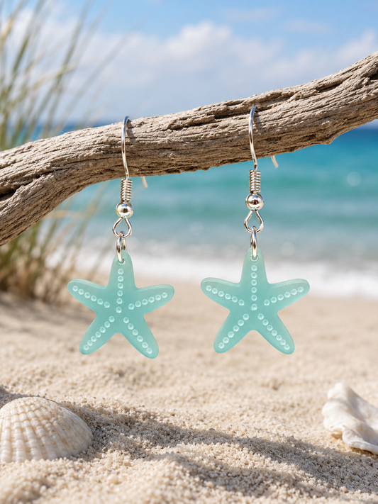 A pair of starfish-shaped dangling earrings in pale mint green with silver hooks, hanging from a driftwood branch on a sandy beach with the ocean and sky blurred in the background.