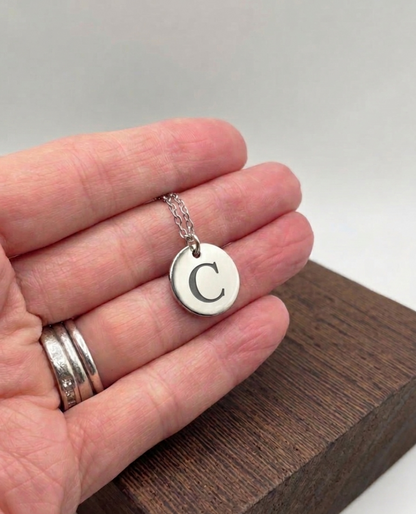 Silver necklace with a round pendant featuring the letter 'C' held in a hand on a wooden surface.